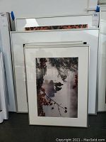 Photo showing multiple framed art pieces stacked against a wall, includes misty outdoor scene with tree branches and figures visible