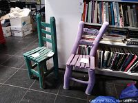 Two painted clay kid-sized chairs with a rough, handmade texture. One greenish-blue, the other lavender, both with ladder-back and slatted seats.