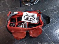 Top-down view of the red canvas camera bag filled with camera and accessories showing multiple compartments and the instruction manual labeled 'Nikkormat FT2'