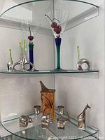 Glass cabinet shelf displaying two martini glasses with green stems, two silver-toned round toothpick holders with green picks, two silver multi-function corkscrews