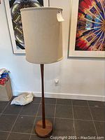 Full view of the lamp showing the wooden base, stand, and the white fabric shade in a room with tiled floor and framed pictures on the wall.