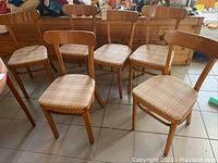 Six matching wooden dining chairs with curved backs and beige plaid upholstered cushions, arranged in two rows on a tiled floor