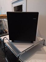 Front and side view of the black cube-shaped Cambridge Soundworks BassCube 10 subwoofer placed on a metal cart, showing its size and shape.