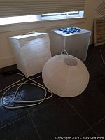 Photo showing two white paper lamps on floor next to a frosted acrylic stand with glass tic-tac-toe on top near a window.