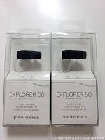 Front view of two new Plantronics Explorer 50 Bluetooth headsets in clear packaging with black earpieces and clear ear hooks.
