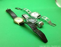 Three wristwatches arranged on green background: Clebar gold-tone with leather strap, Hamilton stainless-tone with perforated bracelet, and silver-tone watch with black dial and expansion band.