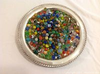 Top-down photo showing hundreds of colorful vintage marbles in a round silver tray with decorative beaded edge.