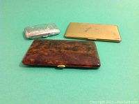 Three cigarette cases displayed on a green background: one silver engraved, one gold-toned, and one faux tortoise shell.