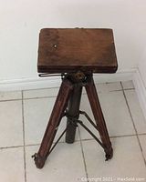 Front view of antique wooden camera tripod with three legs and adjustable top platform, placed on tiled floor.