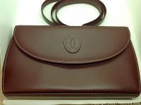 Front exterior view of burgundy leather purse with oval embossed Cartier logo and attached strap laid behind