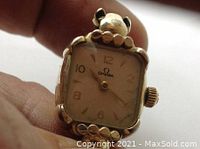 Front view of watch held between fingers showing square dial, Omega logo, applied Arabic numerals and decorative case bezel.