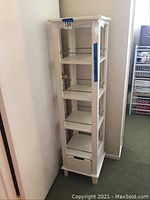 Photo of the white shelving unit showing its tall, narrow structure with four open shelves and a closed drawer at the bottom.
