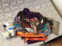 Pile of assorted scarves with multiple colors and patterns folded on table, some scarves have fringe edges.