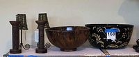 Two carved wooden candle holders with metal chain fringe and two decorative bowls, one wooden and one black with floral design, displayed on a shelf.
