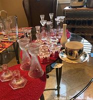 Glass and crystal candlestick holders arranged on red fabric on a glass table, including some paired sets and a teacup with saucer.