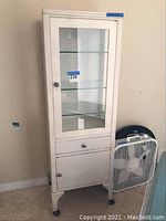 Front view of painted white metal cabinet with glass panels, caster wheels visible, and adjacent fan for scale