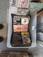 Top view of the plastic container holding the open black CD binder with CDs inside, showing various music CDs including one labeled Decca.