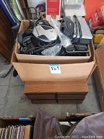 Box containing various electronic items including a vintage Verizon cordless phone, chargers, a portable radio, and VHS tapes stored inside a box.