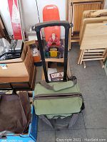 Photo showing a green cooler cart with telescoping handle, surrounded by boxes and other items on floor