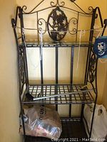 Full view of the black metal shelving unit with glass top and decorative grapevine motif on side panels and back. Shelf folds.