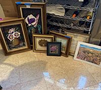 Six framed art pieces arranged on floor showing variety of floral and landscape images in gold and white frames