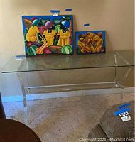 Glass side table with clear acrylic legs, shown with colorful paintings on and behind it for scale reference.