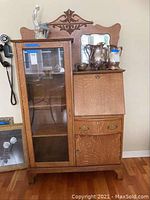Front view of vintage wood secretary desk with glass display case on left, drop-front writing desk, drawer, and cabinet door on bottom right.