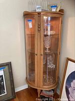Front view of wood and glass lighted locking display cabinet with glass shelves inside and various glassware displayed.