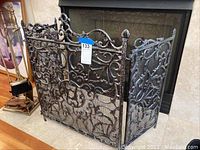 Front and side view showing full three-panel ornate metal fireplace screen with mesh backing in front of a fireplace, metal scrollwork patterns prominently visible.