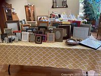 Large assortment of picture frames displayed on a table in various styles, sizes, and colors including ornate gold, wood, and plain frames.