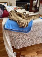 Various table runners and tablecloths folded and stacked on a table, including beige lacework tablecloth covering the table, blue runner with white stitch border, and beige runner with tassels on top.