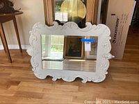 Front view of the white decorative mirror showing ornate metal frame and beveled mirror surface reflecting room interior.