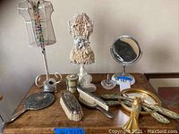 Photo showing vintage brushes, ring holder, table mirror, jewelry holder, decorative metal dress form, and Art Nouveau hand mirror on a wooden surface.