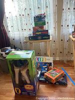Multiple boxed board games stacked on a wooden tray table with 'Life' game and Halloween costume box nearby on floor under a window with sheer curtains.