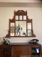 Front view of the Eastlake style wooden mirror with detailed carvings, multiple mirror sections, and shelves on either side. The mirror hangs above a wooden dresser with antique rotary phones on it.