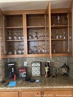 Wine glasses of various styles inside wooden cabinet and several bar items on granite counter