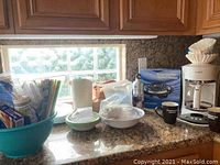 Kitchen countertop with Mr. Coffee coffee maker, portable burner box, ceramic mugs, water bottles, plastic plates and bowls, disposable utensils and cups organized near a window.