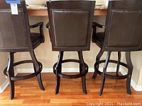 Rear view of three black faux leather rotating bar stools with wooden frames and armrests placed at kitchen counter