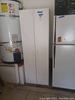 Tall white storage cabinet with two doors, situated next to a water heater and refrigerator.