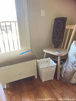 Wooden bench with beige cushion, light wood chair, white decorated waste basket, and South American carved wooden mask resting on chair