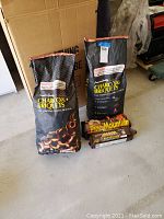 Two black 20lb bags of Pine Mountain Charcoal Briquets with two brown Pine Mountain Crackling Hearth Logs in front on a concrete floor.