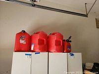 Three 6-gallon and one smaller red gasoline containers seen on white cabinets