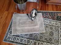 Small beige rug with rectangular design and electric iron placed on top, on a larger floral patterned rug.