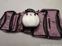 Six soft-sided purple and black bags and one white round hard-sided bag laid out flat