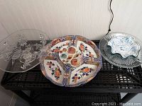 Clear glass cake stand with raised fruit design on bottom, porcelain divided lazy Susan with floral design, and glass veggies and dip plate on the side