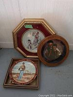 Two framed Norman Rockwell plates and one Buddy Holly commemorative plate on floor against wall.