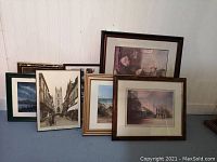 Six assorted framed prints arranged against wall including a framed Norman Rockwell print, two winter landscape prints, and various historic street and interior scenes.
