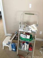 Step ladder with items on shelf and floor beside it including a white step stool and extension cord box