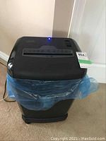 Front angle of the black Staples paper shredder with a blue plastic liner inside the bin. Shredder is plug connected to power.