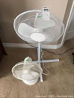 Smaller tabletop fan in front and larger pedestal fan behind, both white, on floor near wall outlet